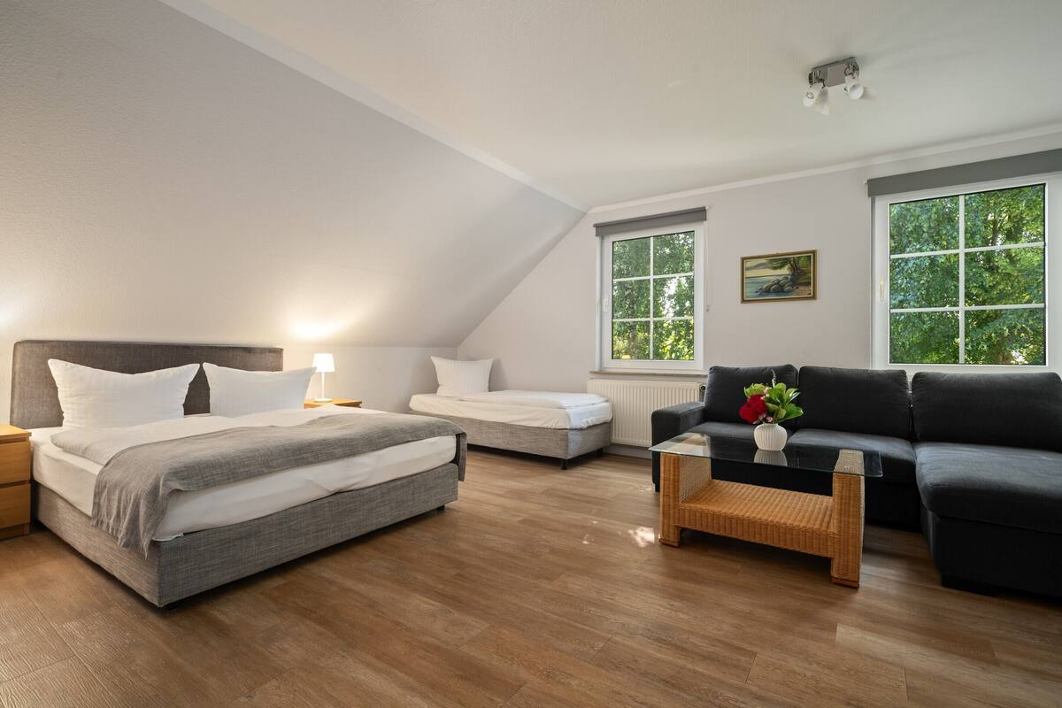 Un dormitorio moderno con una cama grande, una cama más pequeña, un sofá y una mesa de centro. Hay dos ventanas con vistas a árboles.