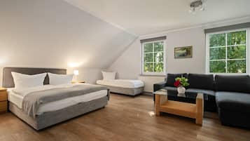 A modern bedroom with a large bed, a smaller bed, a sofa, and a coffee table. There are two windows with views of trees.