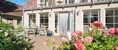 Een gebouw met twee verdiepingen, een balkon, een terras met tuinmeubilair en een tuin met roze bloemen.