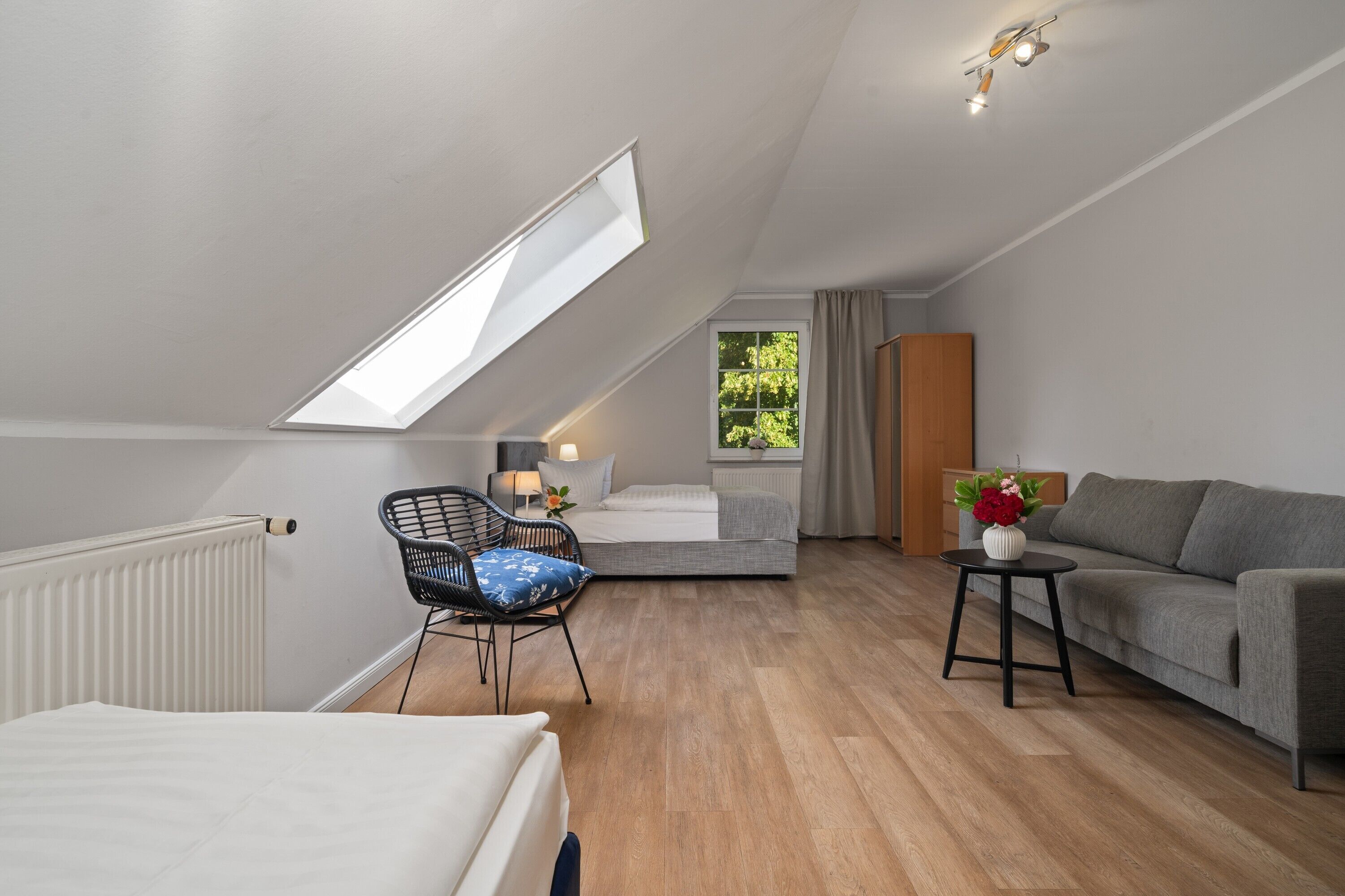 A modern, well-lit room with a sloped ceiling, a bed, a sofa, a chair, and a small table with a vase.