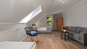 Una habitación moderna y luminosa con techo inclinado, cama, sofá, sillón y una pequeña mesa con un jarrón.