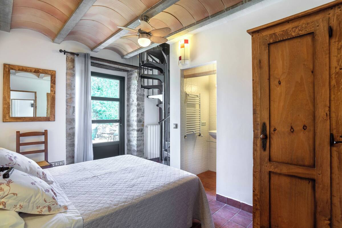 A bedroom with a bed, a wooden wardrobe, a spiral staircase, and a bathroom with a sink and towel rack.