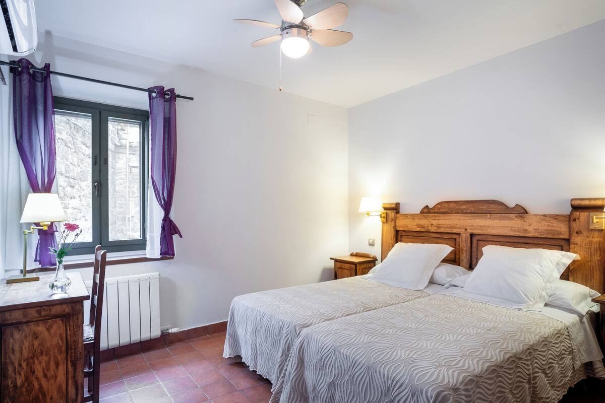 A bedroom with a bed, a desk with a lamp, a window with purple curtains, a radiator, and a ceiling fan.