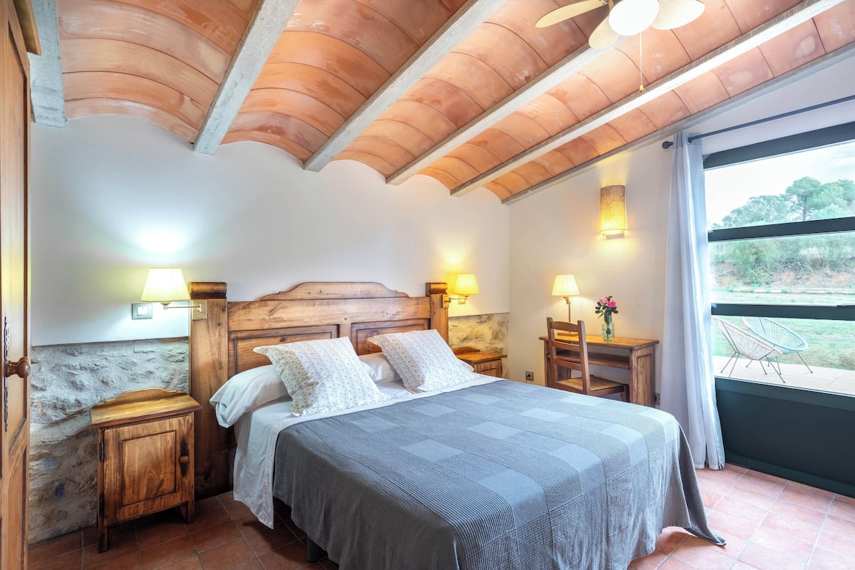 A bedroom with a wooden headboard, two bedside tables with lamps, a large bed with a grey bedspread, and a view of the outdoors through a window.