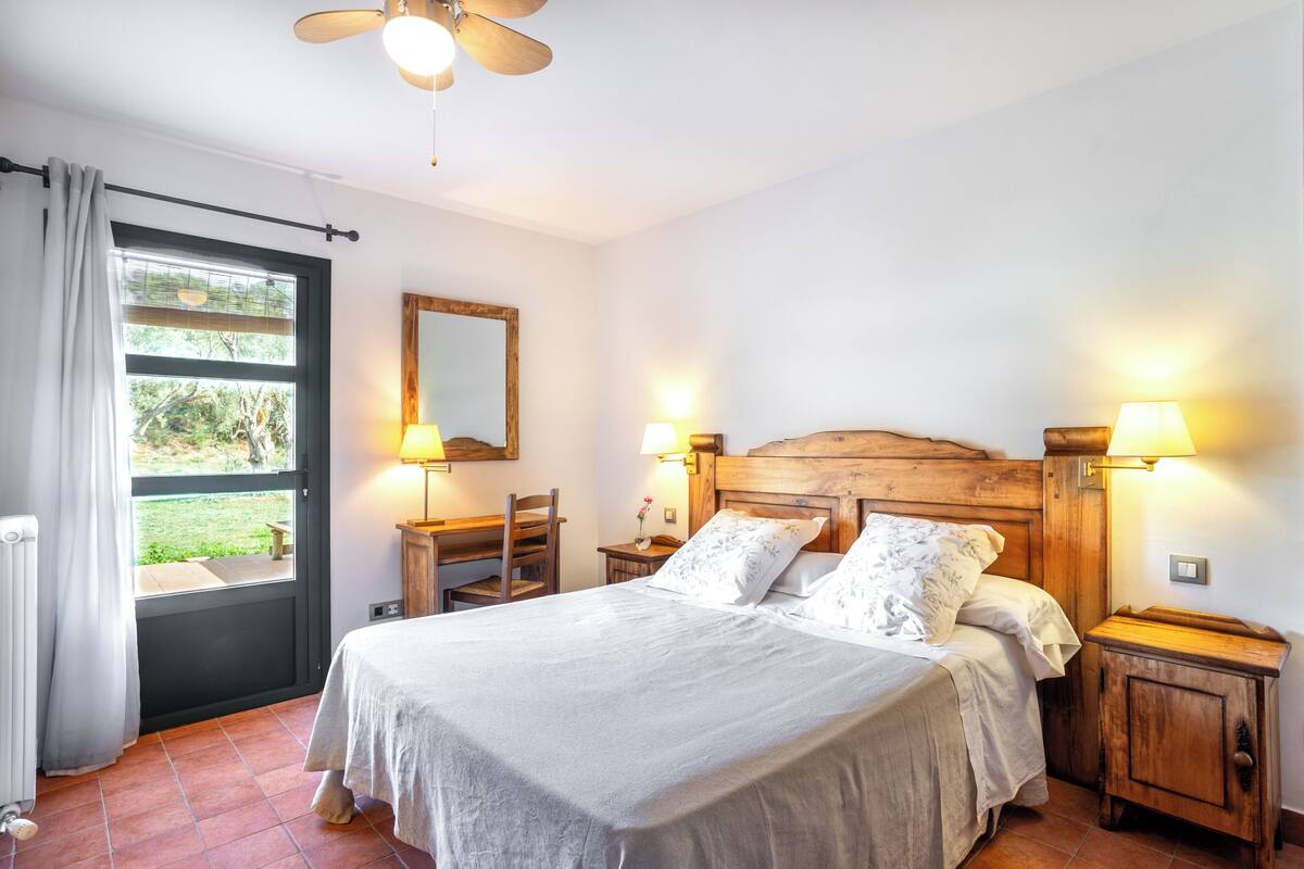 A bedroom with a bed, two bedside tables with lamps, a wooden headboard, a window with curtains, and a view of greenery outside.