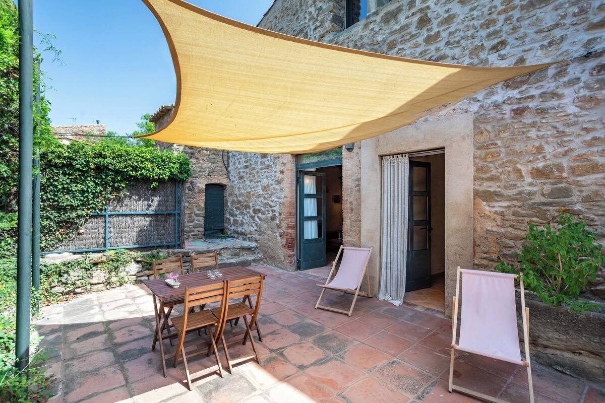 A stone building with a patio area featuring a wooden table and chairs, a canvas shade, and a small easel.