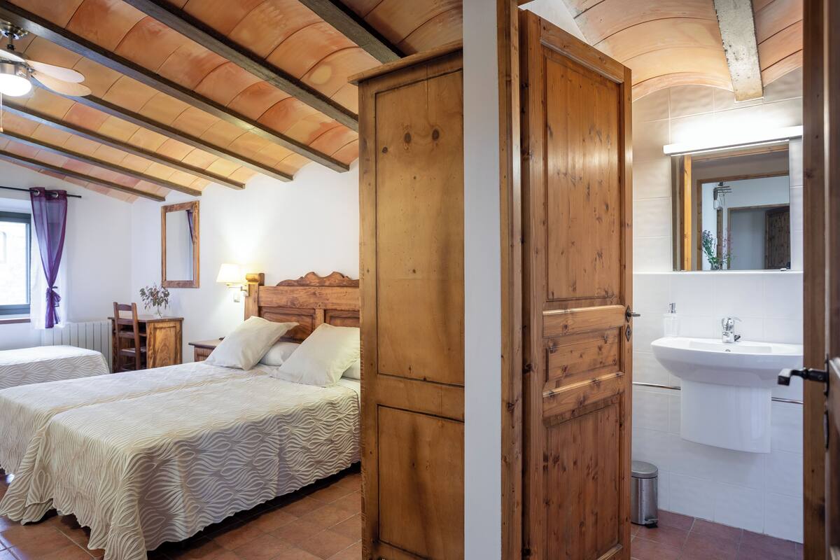 A bedroom with two beds, wooden ceiling beams, a wooden door, a bathroom with a sink and mirror.