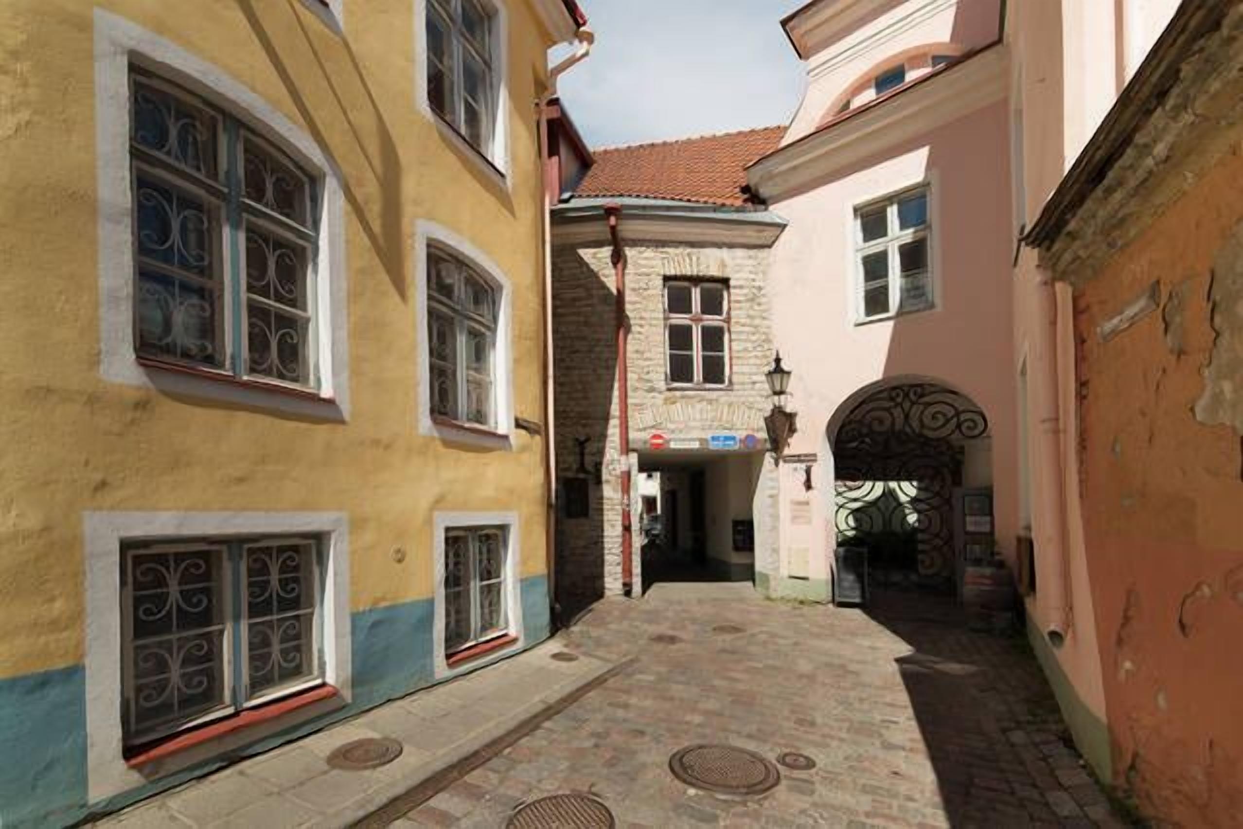Un estrecho callejón adoquinado flanqueado por edificios, con grandes ventanales y rejas de hierro decorativas, y un edificio de piedra con tejado de baldosas al fondo.