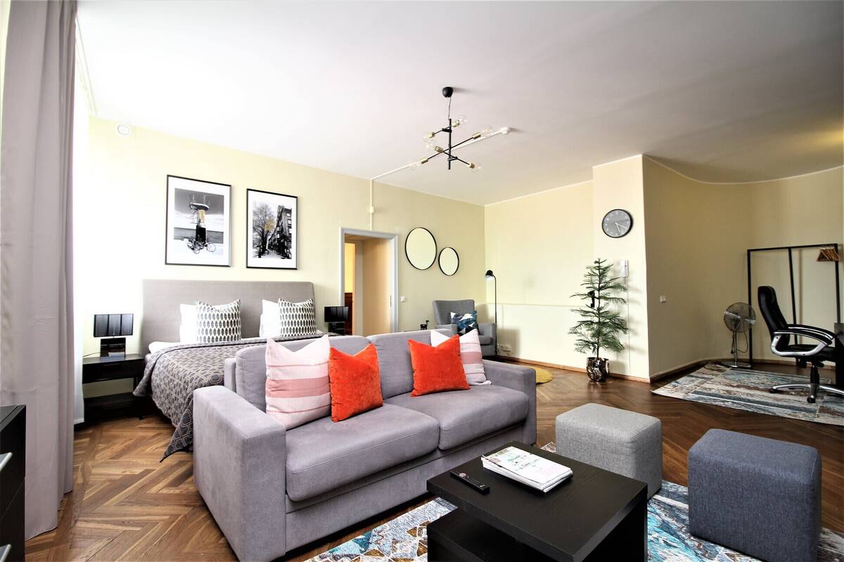Ein modernes Wohnzimmer mit einem grauen Sofa, einem Couchtisch und einem Fernseher. An den Wänden hängen gerahmte Bilder, eine kleine Pflanze steht dort, und es gibt eine Uhr.