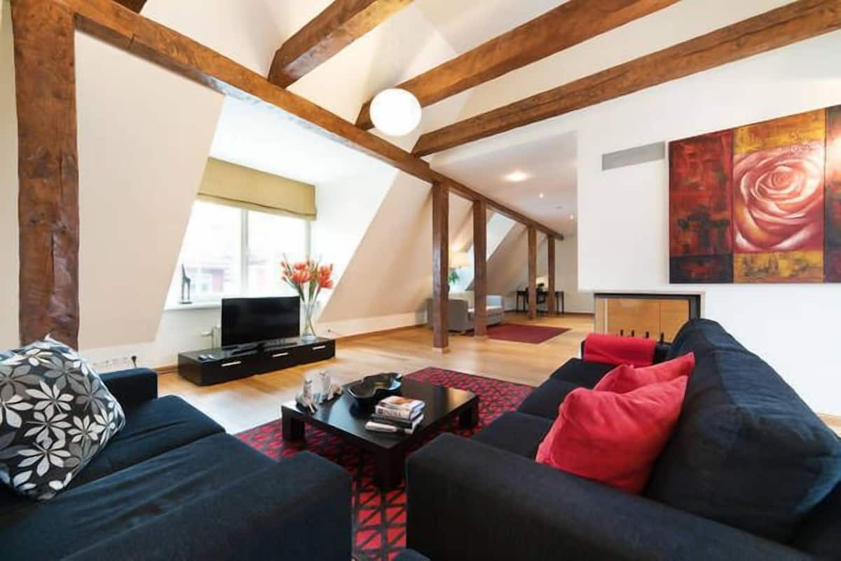 Ein modernes Wohnzimmer mit einer dunklen Couch, einem Couchtisch und einem Fernseher. An der Wand hängt ein großes Gemälde und aus dem Fenster bietet sich ein schöner Ausblick.