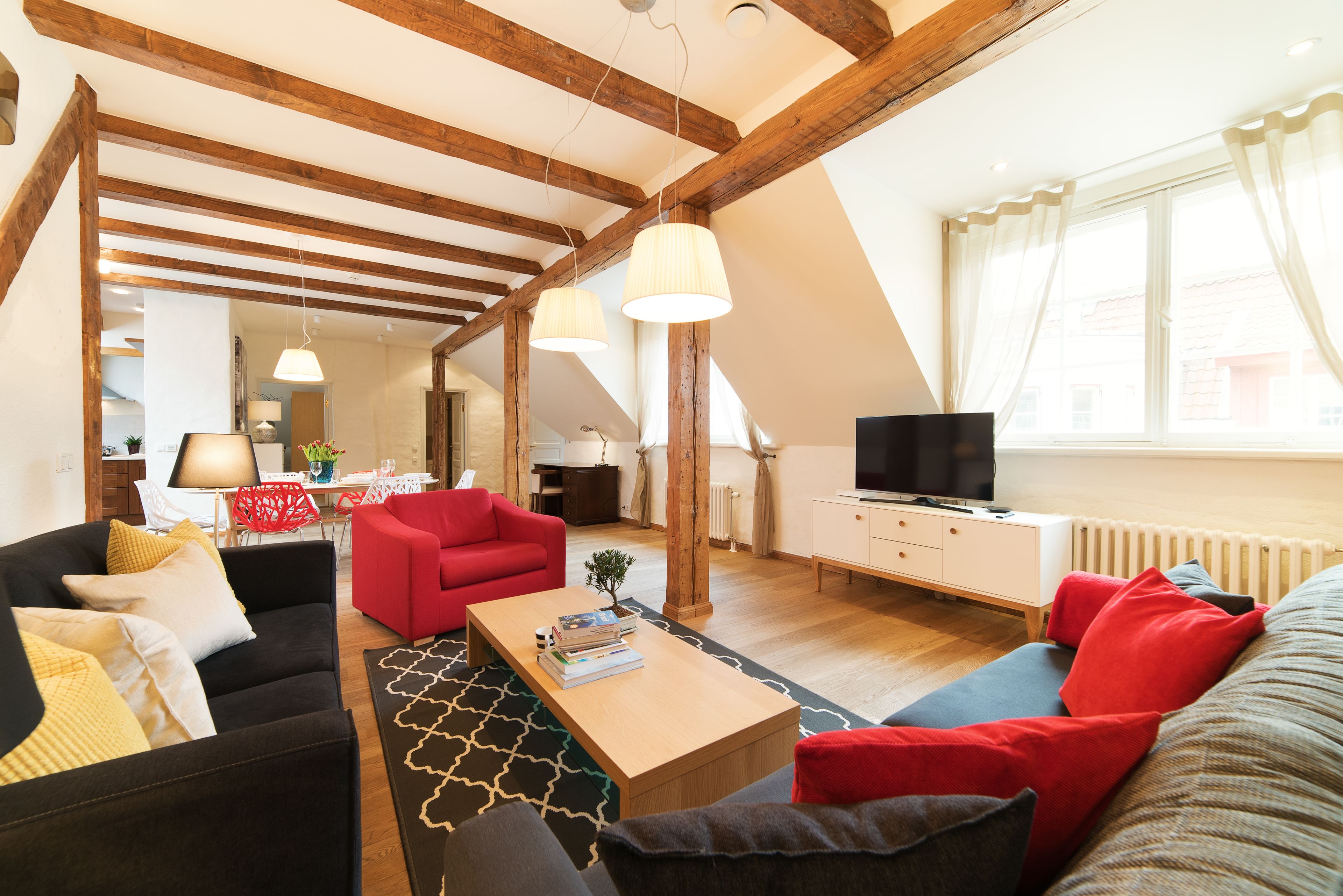 A spacious living room with a high ceiling, wooden beams, a red sofa, a black sofa, and a wooden coffee table.