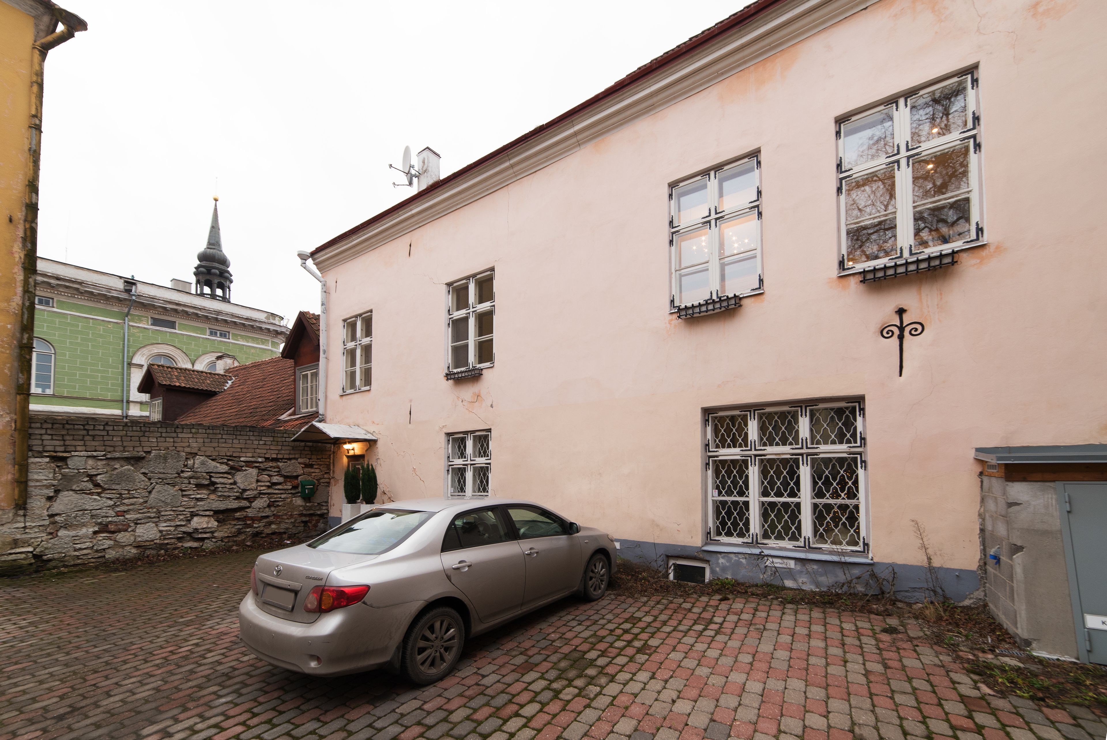 A building with a car parked in front, stone wall, and a church spire in the background.