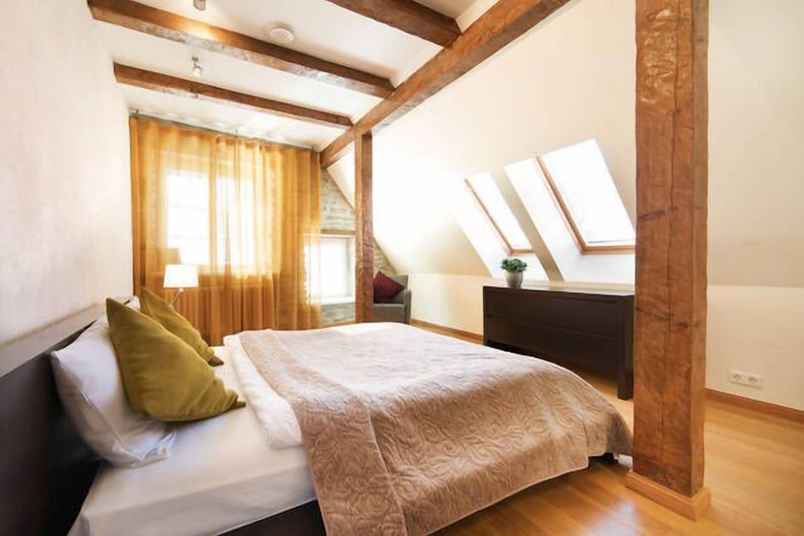 A bedroom with a bed, wooden beams, a window with curtains, a dresser, and a plant.