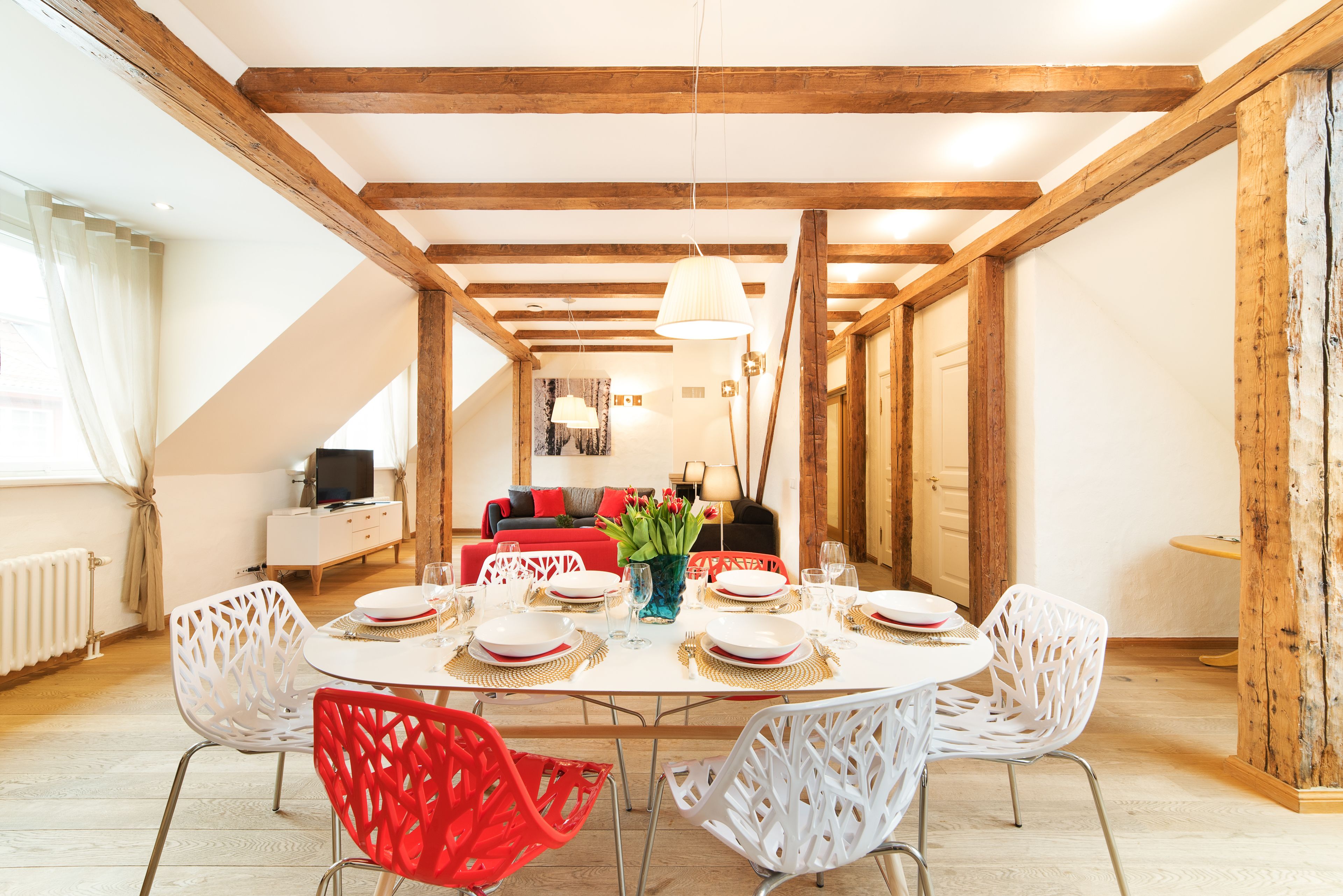 Un comedor con una mesa redonda dispuesta para cuatro personas, sillas blancas y una silla roja. Se ve un televisor al fondo.