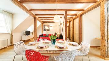 Un comedor con una mesa redonda dispuesta para cuatro personas, sillas blancas y una silla roja. Se ve un televisor al fondo.