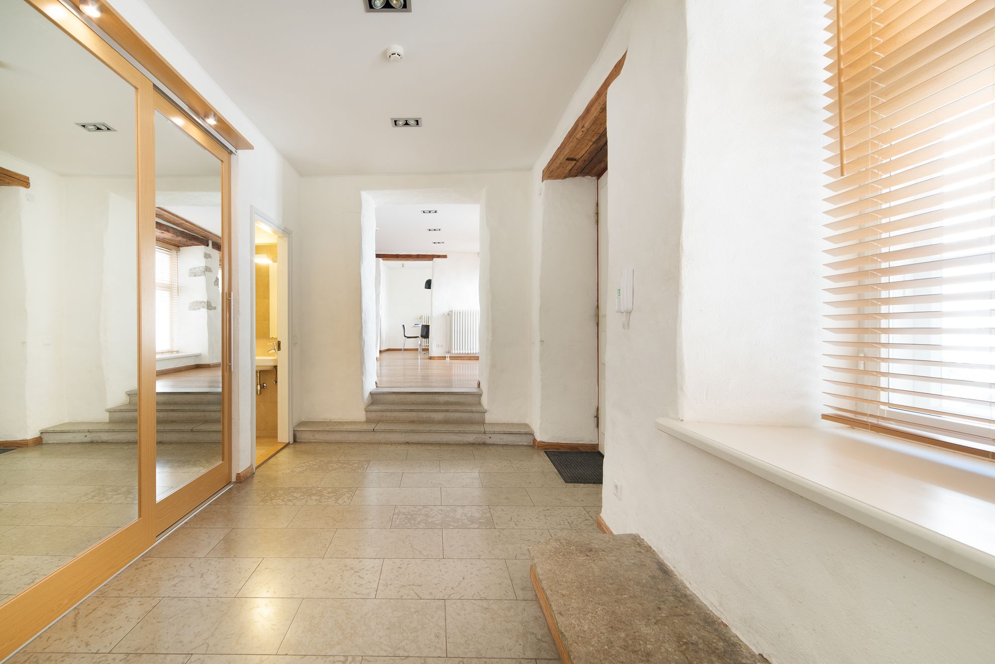 A hallway with a mirrored wall, a wooden floor, and a window with blinds.