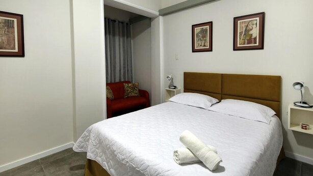 A neatly made bed with a white comforter and a rolled towel, a red chair with a patterned cushion, and two framed artworks on the wall. - MZ Apartments Atlantica posto 6 (Rio de Janeiro)