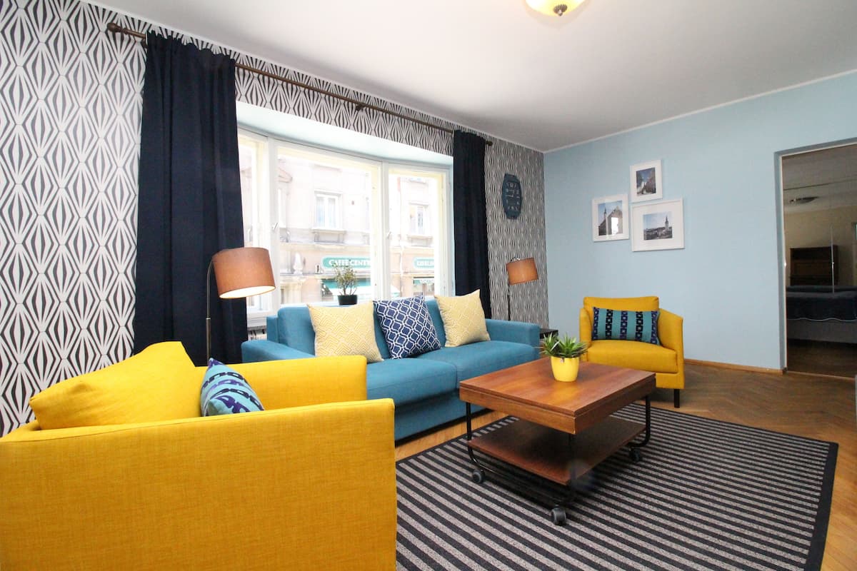 A living room with a blue sofa, yellow armchairs, a wooden coffee table, and patterned curtains.