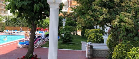 Zona junto a la piscina con un pilar blanco, un árbol y un jardín bien cuidado.