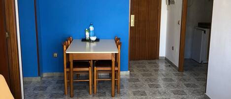 A room with a blue wall, a wooden dining table with chairs, a TV mounted above the table, and a bed with a wooden headboard.