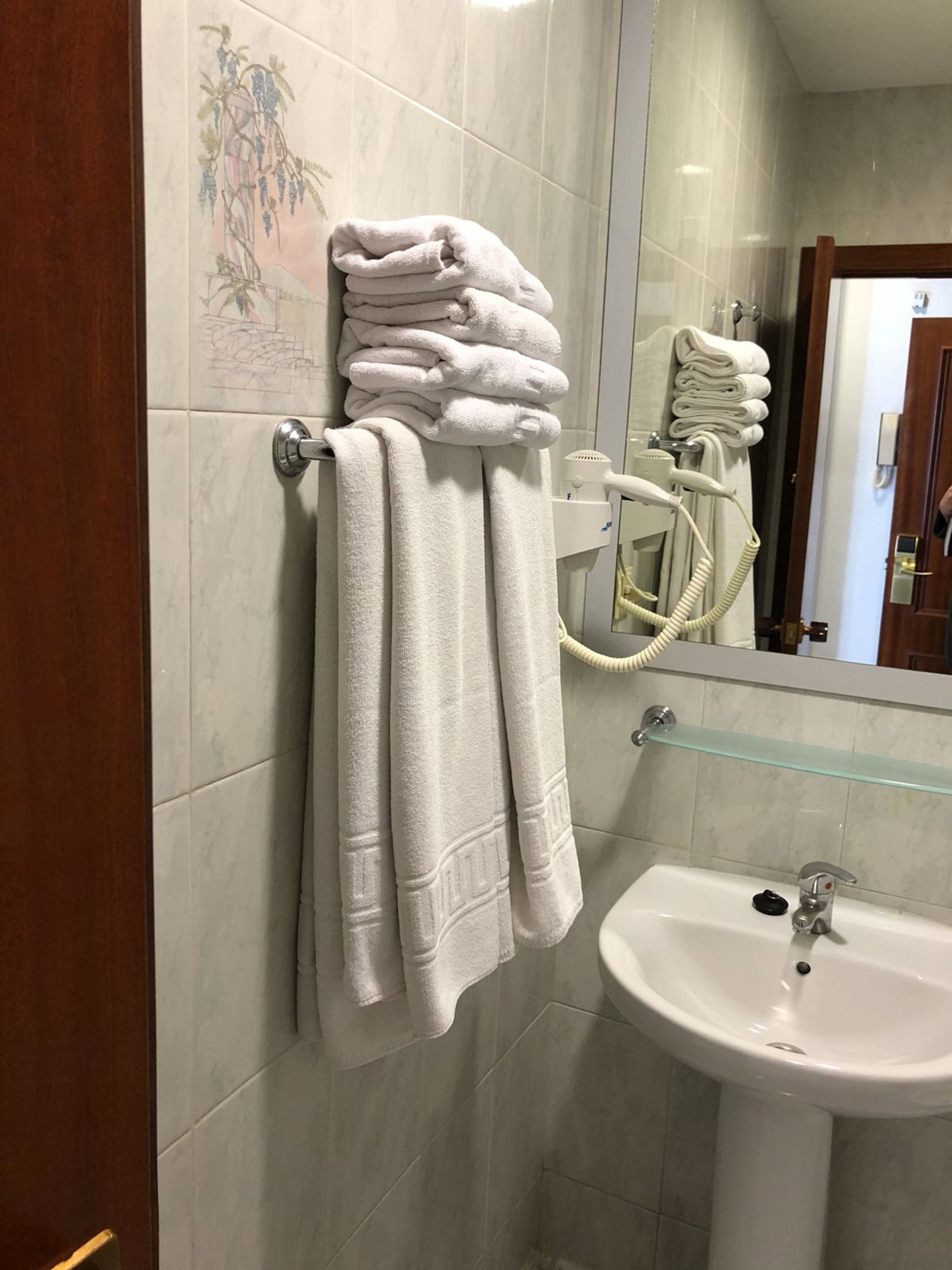 Un baño con una pila de toallas blancas en un perchero, un estante de vidrio sobre un lavabo blanco y un secador de pelo en la pared.