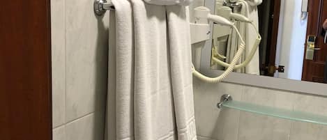 Un baño con una pila de toallas blancas en un perchero, un estante de vidrio sobre un lavabo blanco y un secador de pelo en la pared.