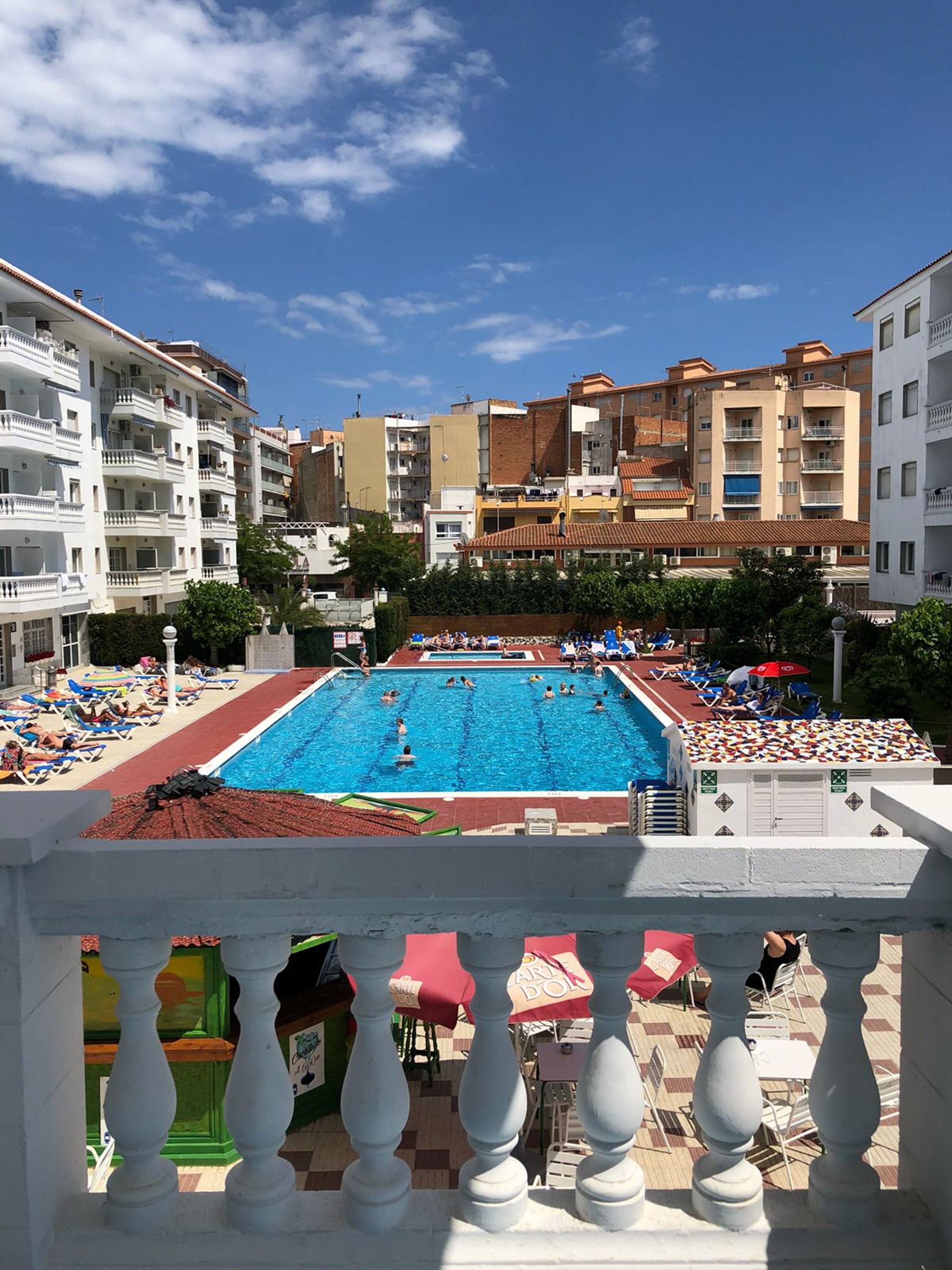 A swimming pool surrounded by multi-story buildings with balconies, a sun terrace with lounge chairs, and a view of the sky.