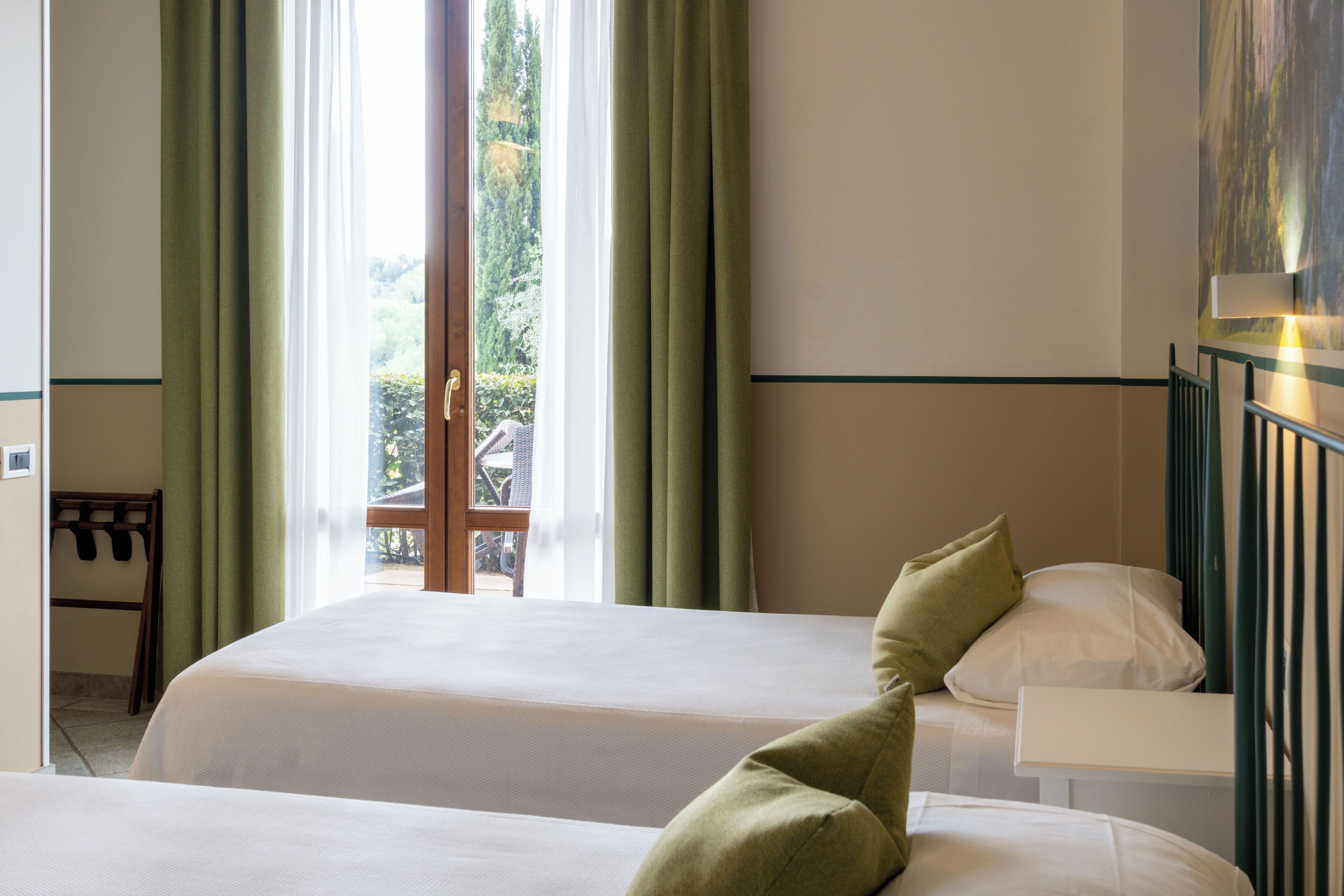A hotel room with two beds, a window with curtains, a view of greenery outside, and a painting on the wall.
