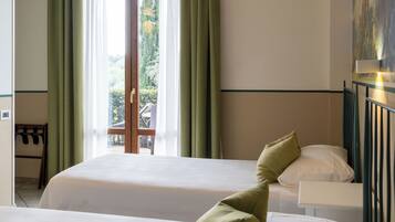 A hotel room with two beds, a window with curtains, a view of greenery outside, and a painting on the wall.