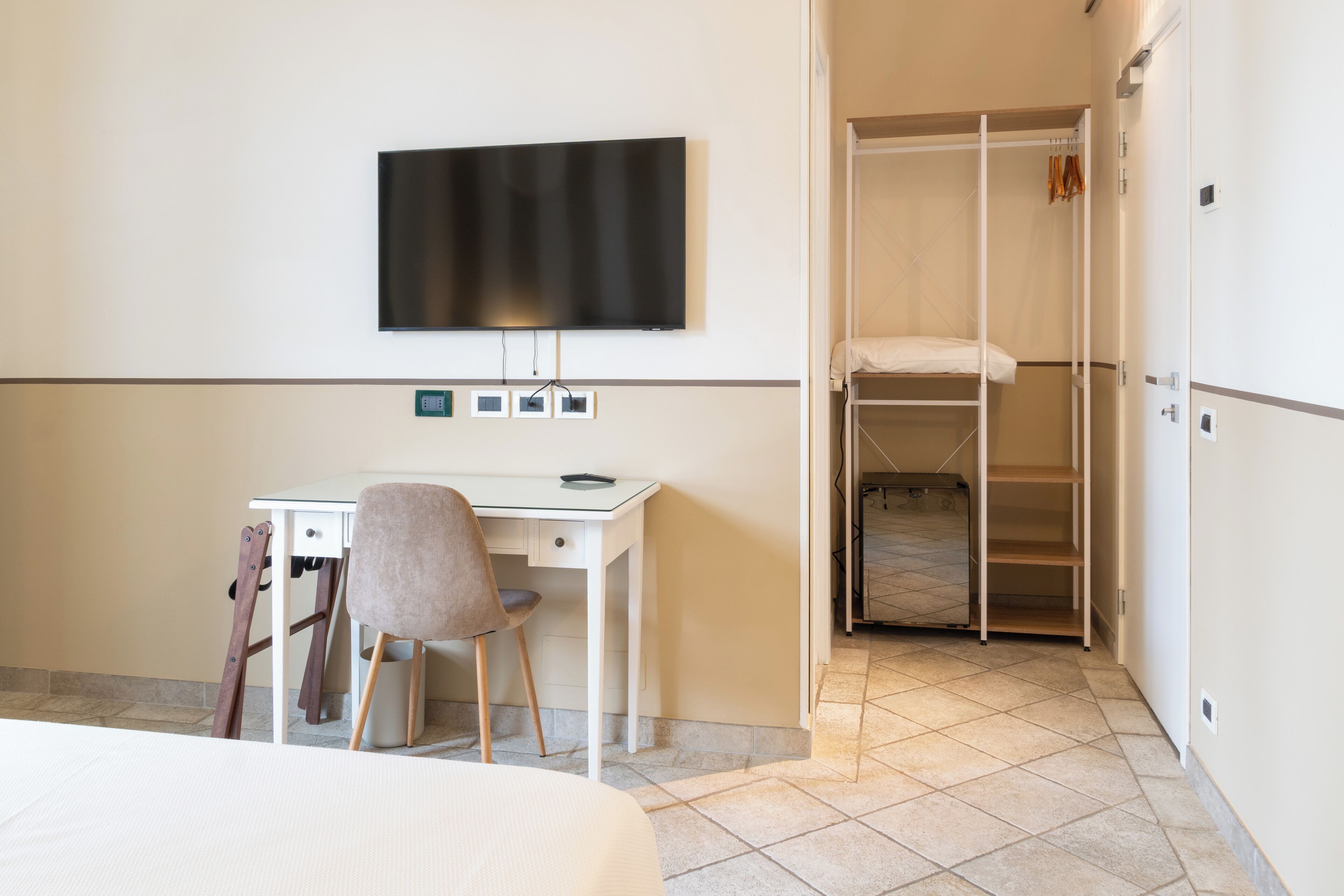Une chambre d’hôtel comprenant un lit, un bureau avec une chaise, un téléviseur fixé au mur et un dressing avec des étagères.
