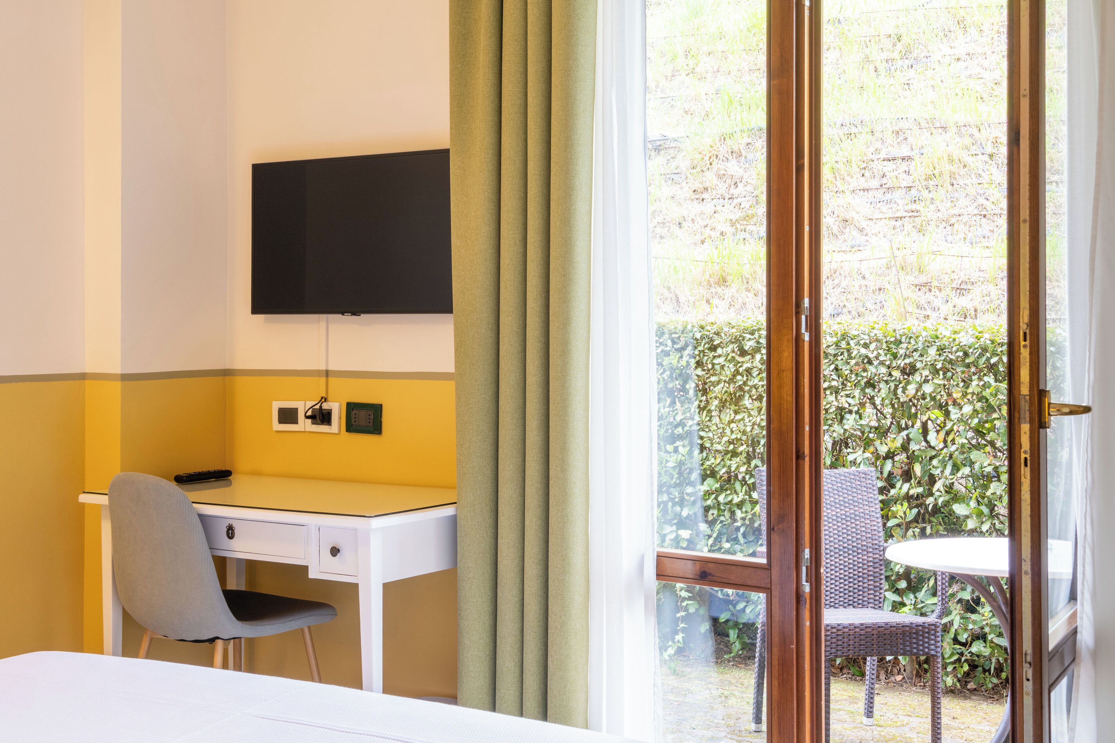 Habitación de hotel con cama, escritorio y silla, televisor montado en la pared y vista a un jardín a través de una puerta de vidrio.