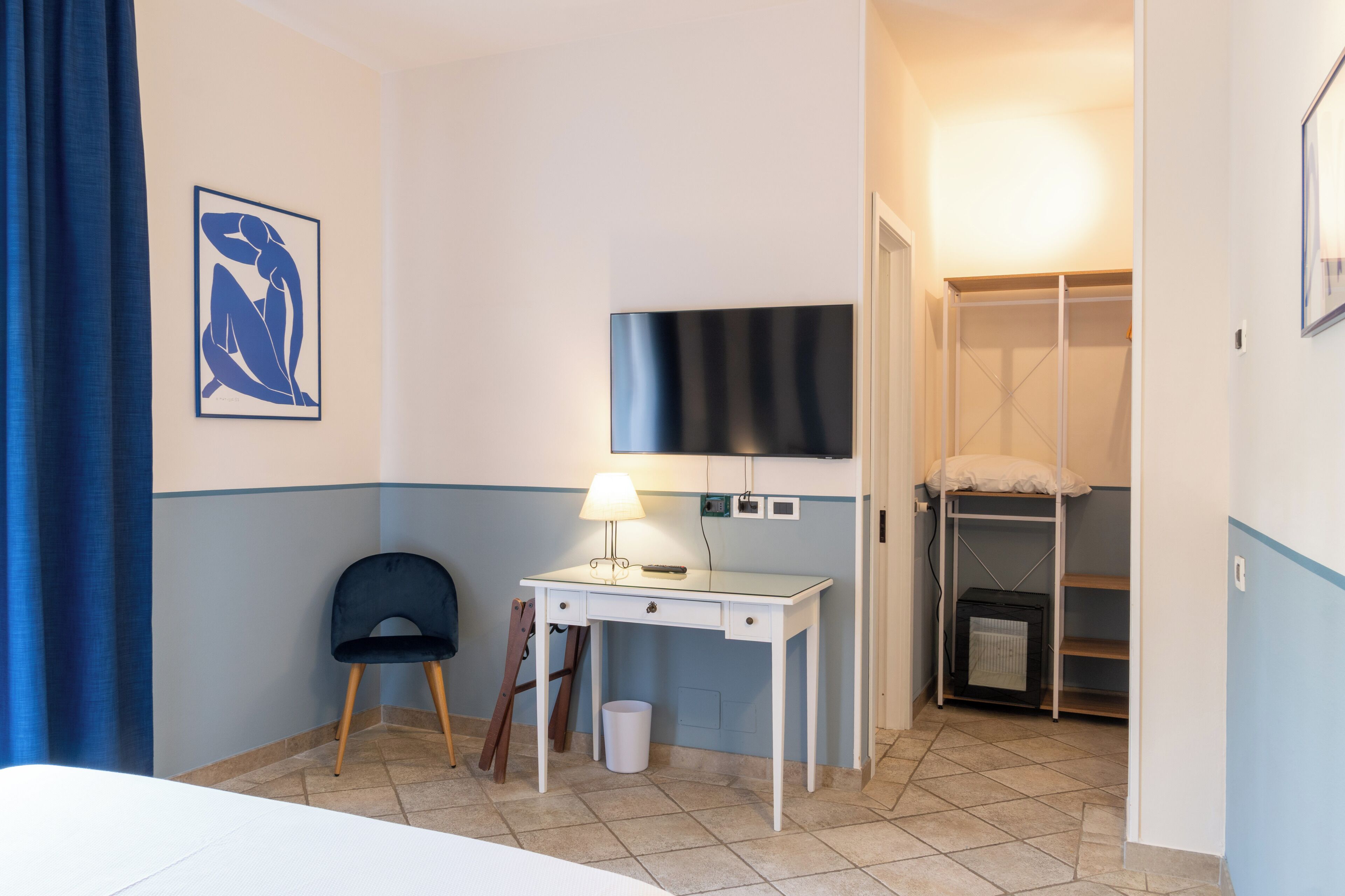 Ein Hotelzimmer mit einem Bett, einem Schreibtisch mit Lampe, einem Sessel, einem an der Wand befestigten Fernseher und einem Schrank mit Ablagefläche.
