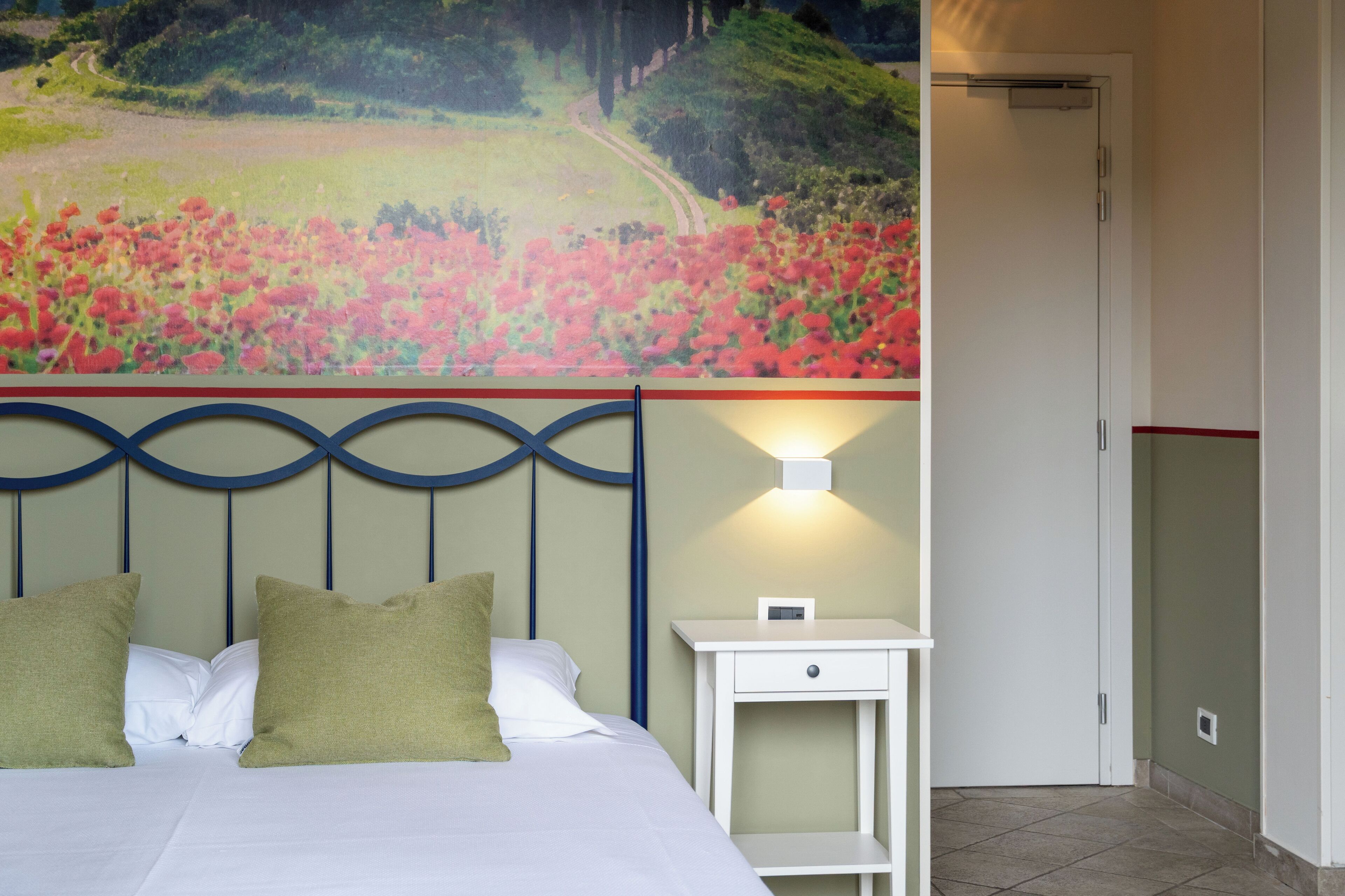 Habitación de hotel con cama, mesita de noche, lámpara de pared y un cuadro grande con un paisaje sobre la cama.