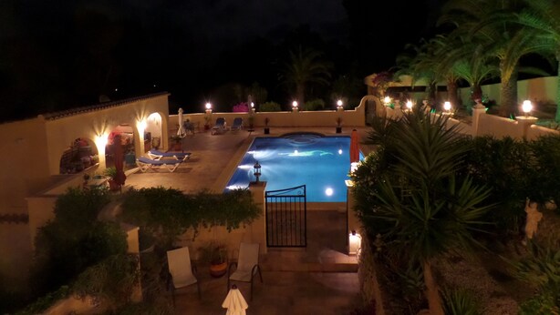 A nighttime view of a pool area with lounge chairs and a gate, surrounded by plants and under a night sky with a visible moon. - Villa Senomar (Benissa)