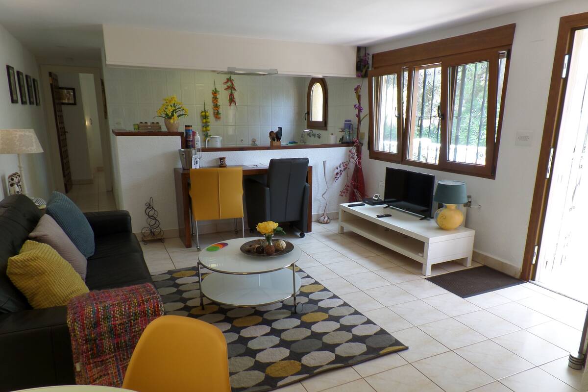 Un salon comprenant un canapé, une table basse et un meuble pour la télévision. Il y a également un coin cuisine avec une fenêtre et une porte donnant sur une autre pièce.