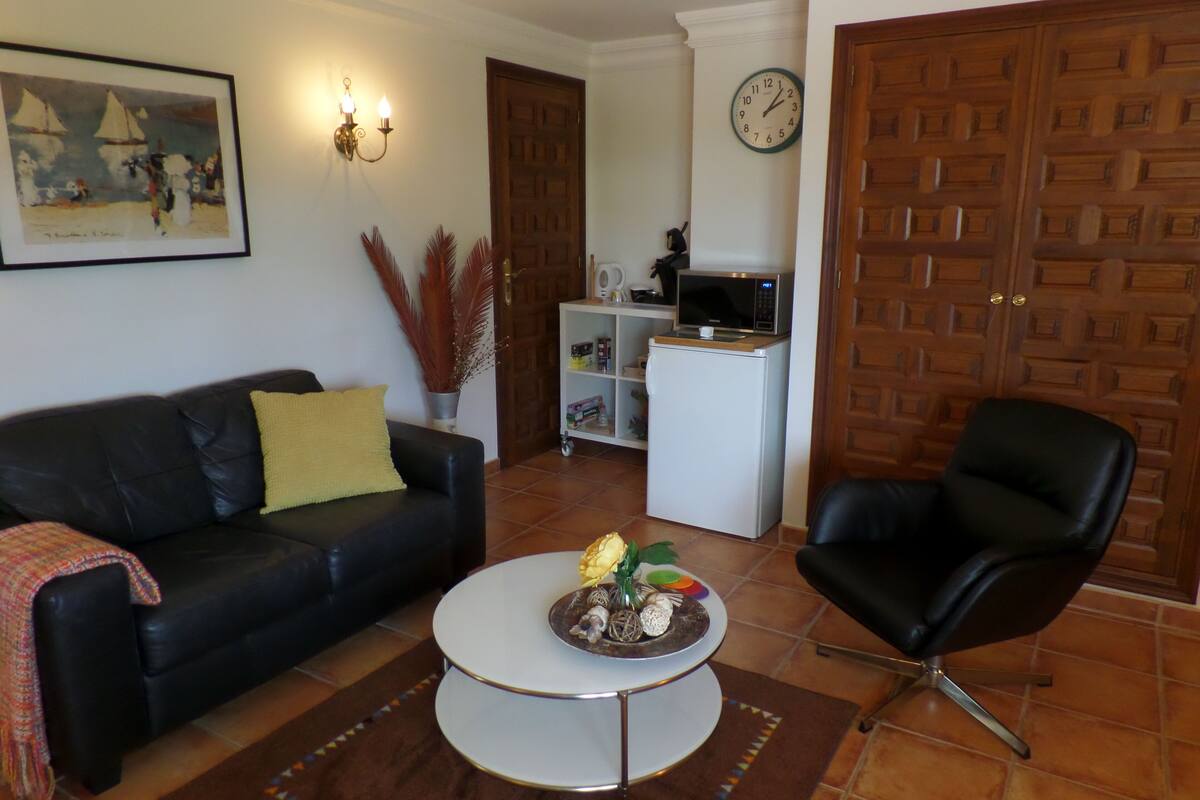 Un salon avec un canapé en cuir noir, une table basse blanche, un fauteuil noir, une horloge au mur et une porte ornée d’un panneau en bois.
