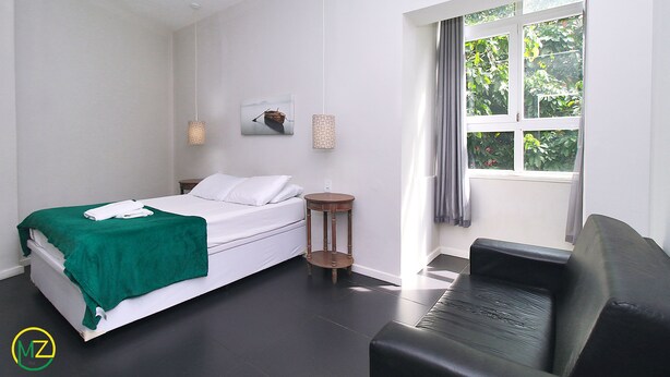 Une chambre à coucher comprenant un lit, une table de chevet, une chaise, une fenêtre avec des rideaux et un appareil de climatisation.