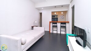 Un salon moderne et minimaliste, avec un canapé blanc, un petit bureau avec un ordinateur et un coin cuisine à l’arrière-plan.