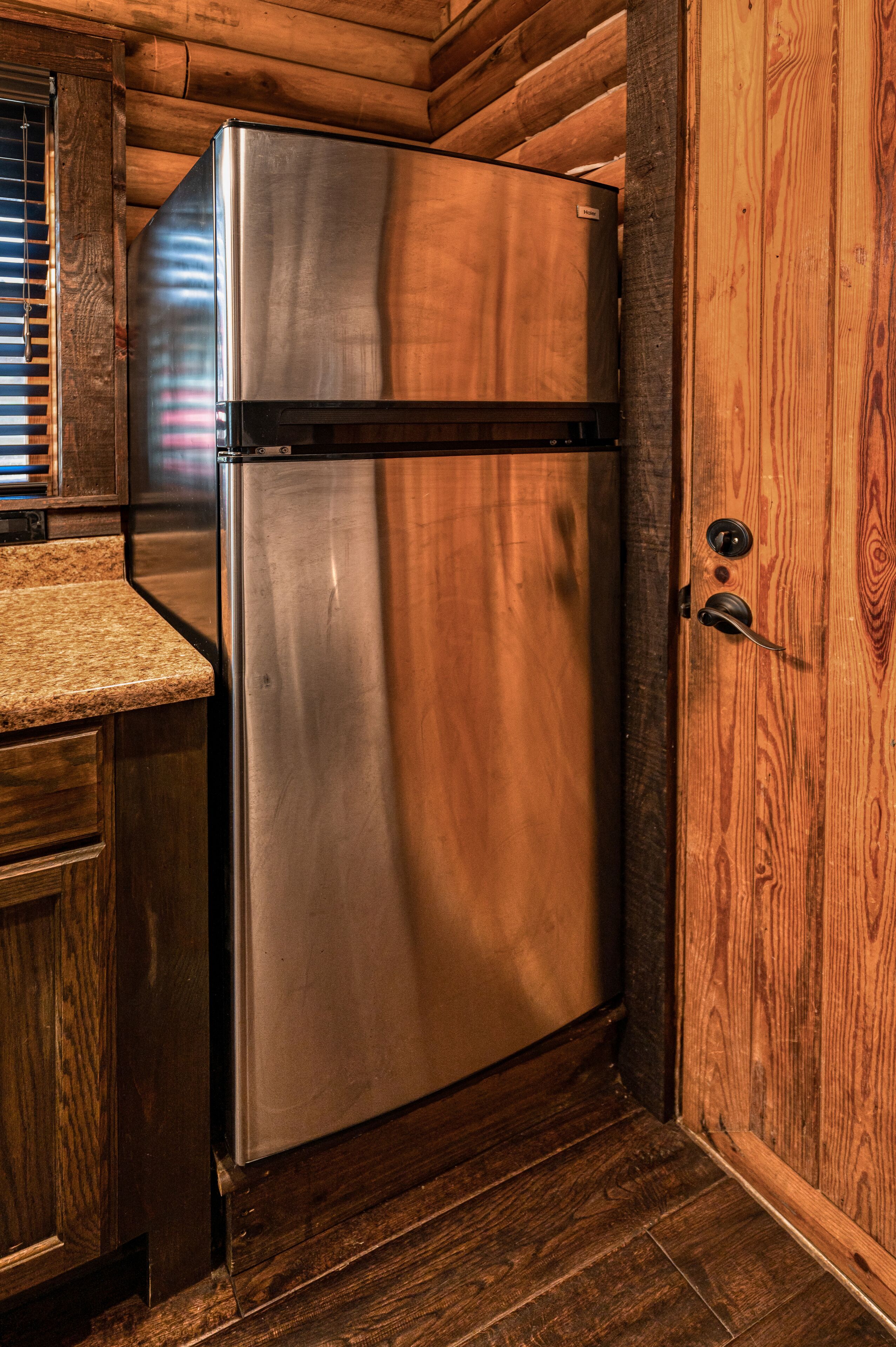 A stainless steel refrigerator in a wooden cabin-style kitchen with a wooden door and a window with blinds.