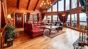 A spacious wooden cabin with a large living area featuring a red leather sofa, a coffee table, and a chandelier. Large windows provide a view of the surrounding landscape. - stayNantahala - Smoky Mountain Cabins (Topton)