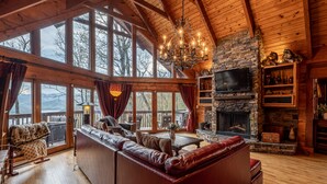 A cozy cabin interior with a stone fireplace, leather seating, and large windows offering a view of the mountains. - stayNantahala - Smoky Mountain Cabins (Topton)