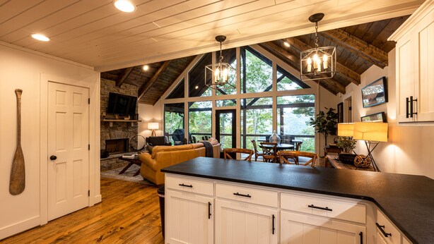 A spacious kitchen with white cabinets, a black countertop, and a stone fireplace. There is a dining area with a table and chairs, and a large window offering a view of the outdoors. - stayNantahala - Smoky Mountain Cabins (Topton)