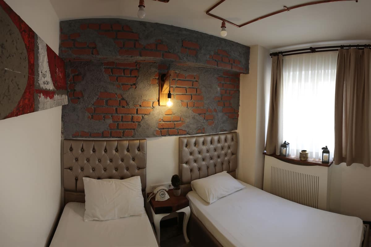 Quarto de hotel com duas camas, parede de tijolos, janela com cortinas, televisão montada e pequena mesa com abajur.