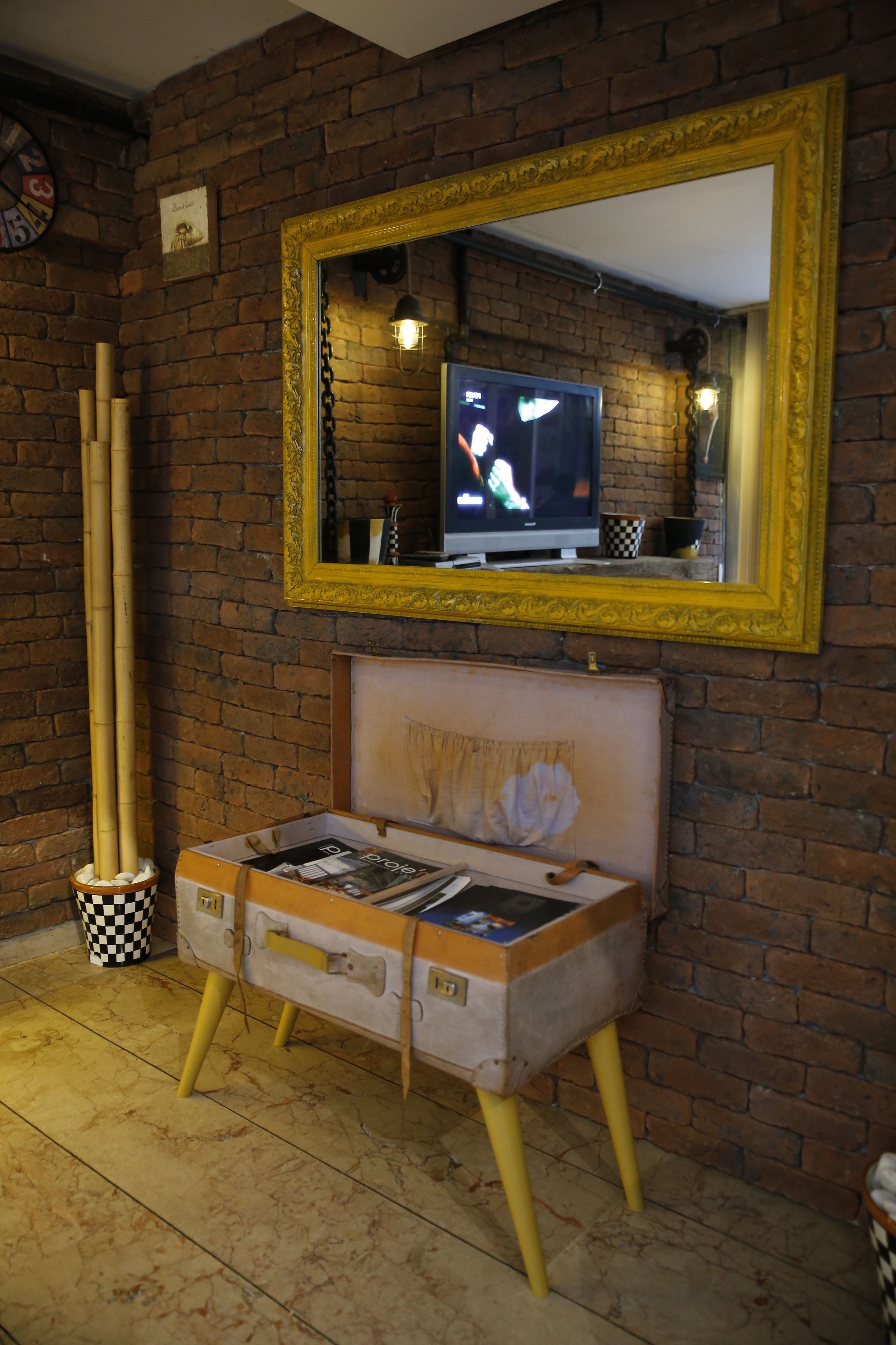 A room with a brick wall, a large mirror, a television, and a vintage suitcase as a seating area.