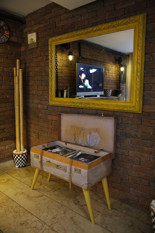 Habitación con pared de ladrillo, un espejo grande, televisor y una maleta antigua utilizada como asiento.