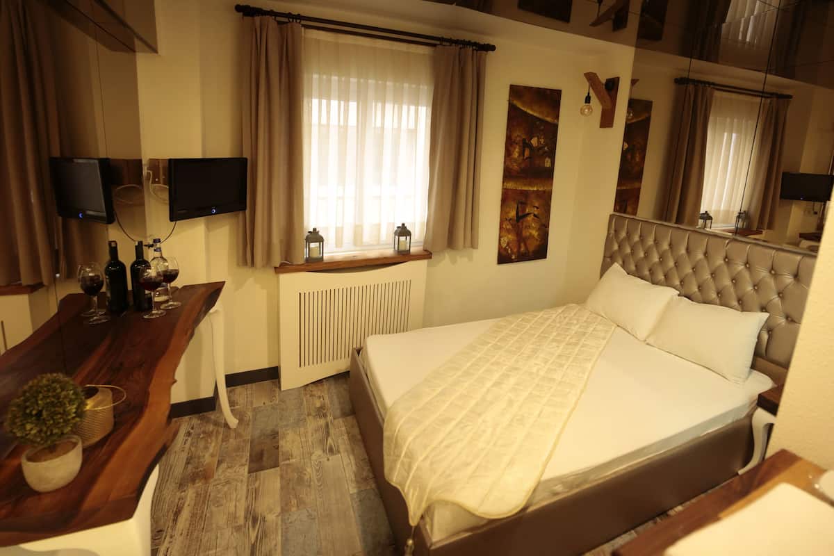 Quarto de hotel com uma cama, uma escrivaninha de madeira com uma planta, televisão e uma janela com cortinas.