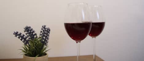 Una planta pequeña en maceta y dos copas de vino sobre una mesa de madera en una habitación con pared blanca y ventana con cortinas.