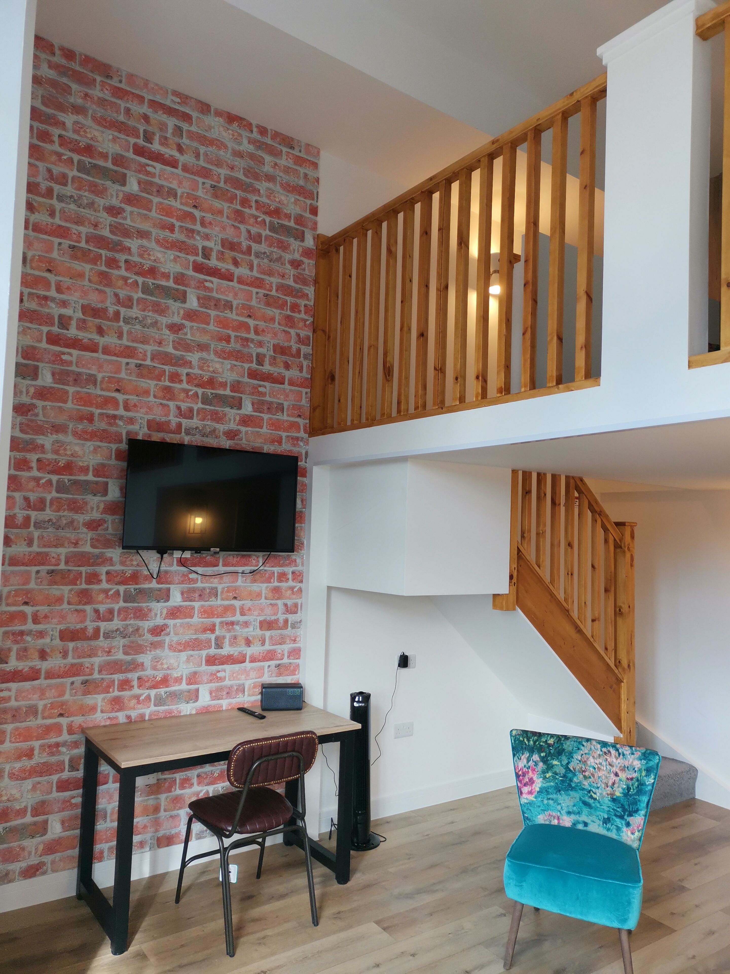 Un cuarto estilo loft con pared de ladrillo, un televisor de pantalla plana, una escalera de madera, un escritorio con una silla y un sillón azul con estampado floral.