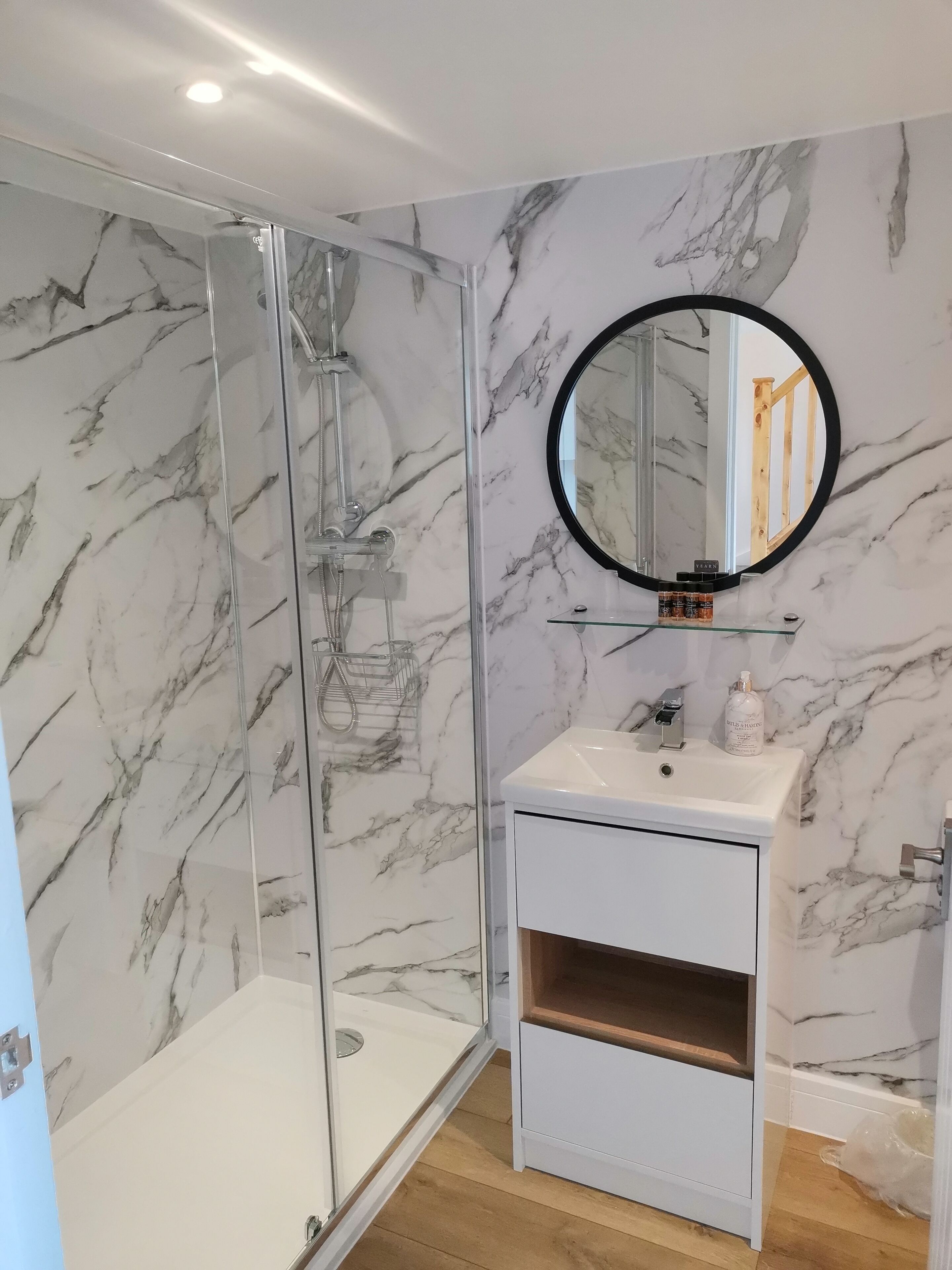 A bathroom with a glass-enclosed shower, a marble-patterned wall, a round mirror, and a white sink with a bottle of shampoo.