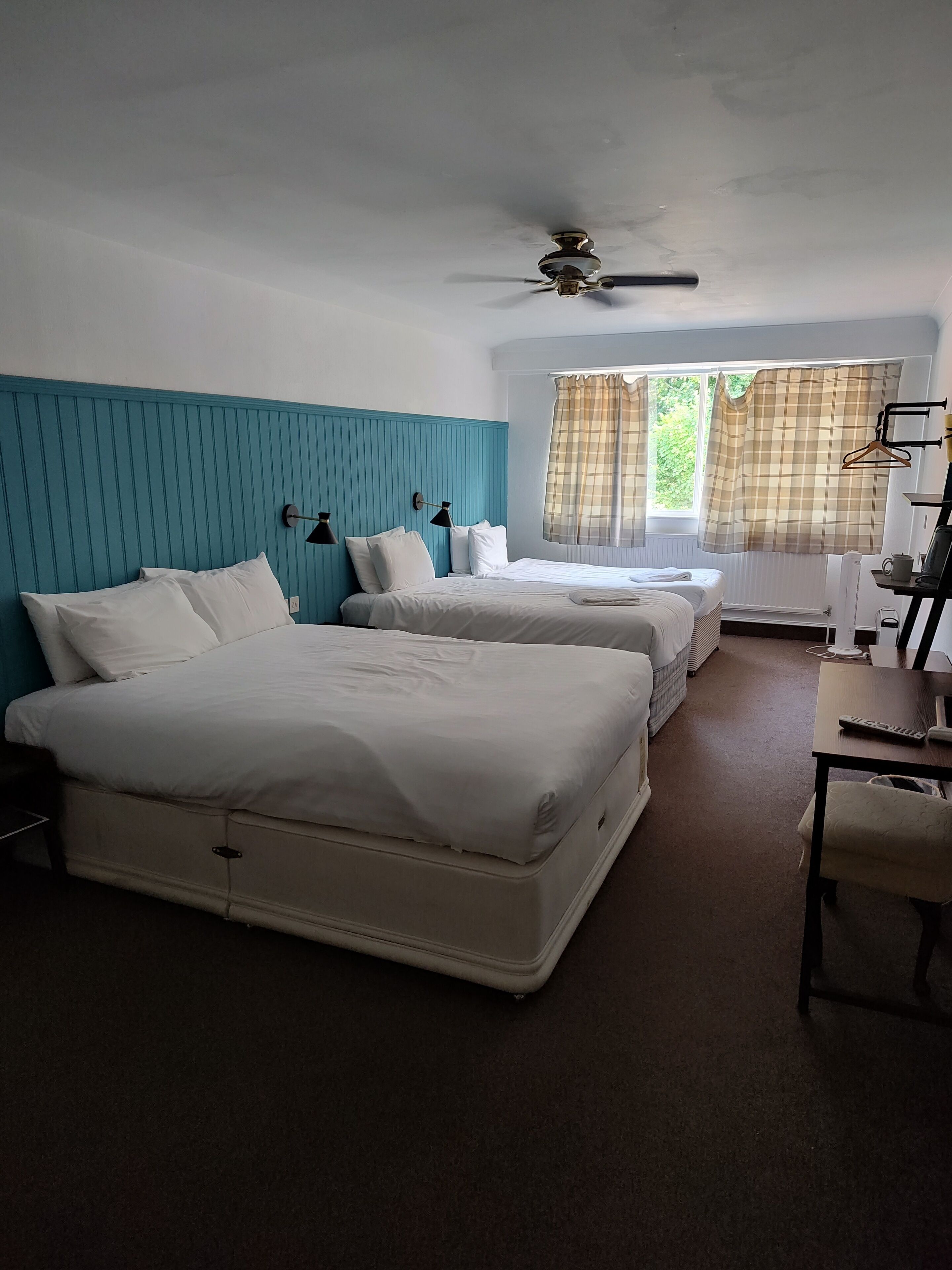 A hotel room with two beds, a desk, a chair, a ceiling fan, and a window with curtains.